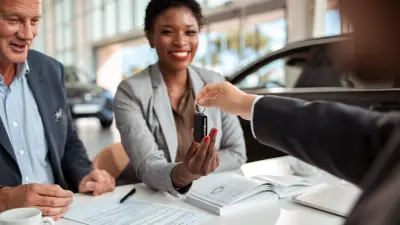 Eine Frau und ein Mann sitzen im Autohaus vor einem Schreibtisch, die Frau nimmt lächelnd einen Fahrzeugschlüssel entgegen.