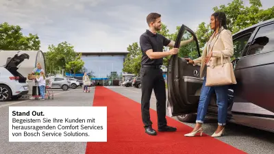 Eine Dame steigt aus ihrem Wagen aus, sie hat eine Tasche über dem Arm hängen. Ein Concierge hält ihr die Tür auf. Die Szene findet auf dem Parkplatz eines Supermarktes statt, auf dem Parkplatz sind weitere Personen zu sehen. 