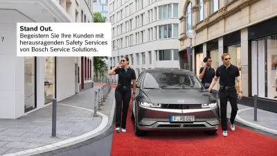 Ein Autofahrer fährt mit seinem Fahrzeug durch eine Straße in einer Innenstadt, er wird von drei Personen begleitet, die aussehen wie Sicherheitsmitarbeiter. Das Fahrzeug fährt auf einem roten Teppich.