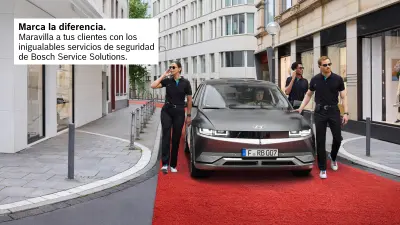 Un conductor conduce su vehículo por una calle del centro de la ciudad, acompañado por tres personas que parecen guardias de seguridad. El vehículo circula sobre una alfombra roja.