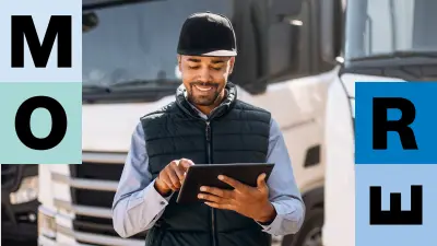 Ein Mitarbeiter eines Logistikunternehmens steht vor verschiedenen Lkw und schaut lächelnd auf sein Tablet. 