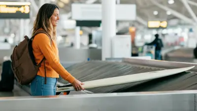 A woman with long hair stands smiling at the baggage carousel at an airport and receives her surfboard.