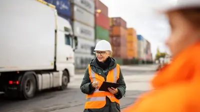 A woman wearing an orange safety vest and a white safety helmet is standing in a logistics terminal, with various containers visible in the background.