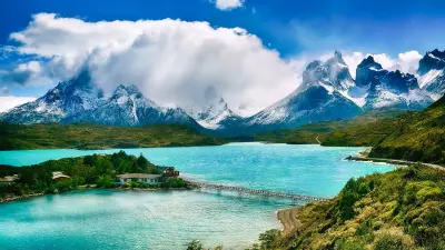 View of a lake in many shades of blue and green, which is surrounded by green meadows. Snow-capped mountains can be seen in the background.