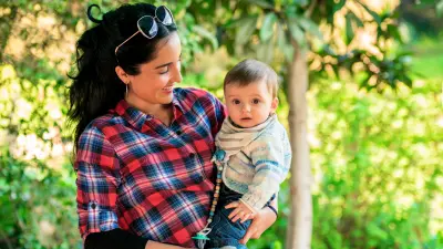 A woman with dark hair looks smiling at a baby she is holding in her arms. She is wearing a red and blue checked shirt and has shoved sunglasses into her hair.