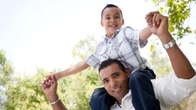 Son is sitting on his father's shoulder, they both smile at the camera