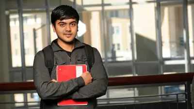A man with dark hair looks at the camera, he has a red portfolio in his hands.