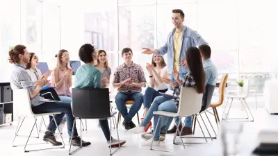 A group of employees sits in a circle, one employee speaks to the group while standing