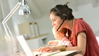 Woman with baby in her arms sits in front of a laptop and types