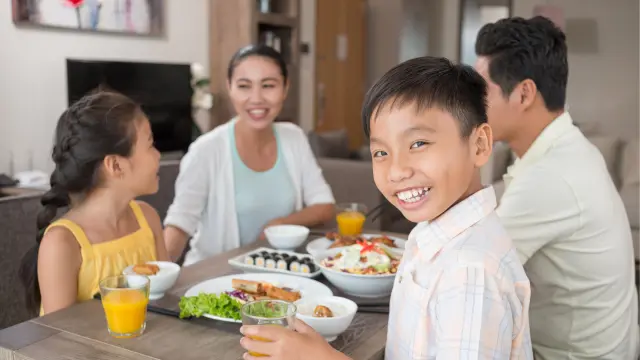 A Vietnamese family is sitting at the kitchen table while eating, mother and son look at the camera smiling.