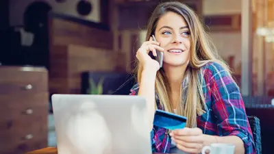 Una mujer está sentada en su escritorio frente a su computadora portátil, hablando por su teléfono inteligente.