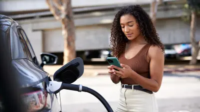 A woman is looking at the smartphone in her hand, she is standing next to her vehicle which is currently being charged electrically.