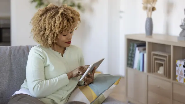 A woman sits on her couch, smiling as she looks at the tablet in her hand.