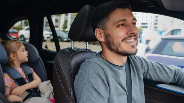 A man is sitting smiling in the driver's seat of his vehicle, with his daughter sitting in a child seat in the back.
