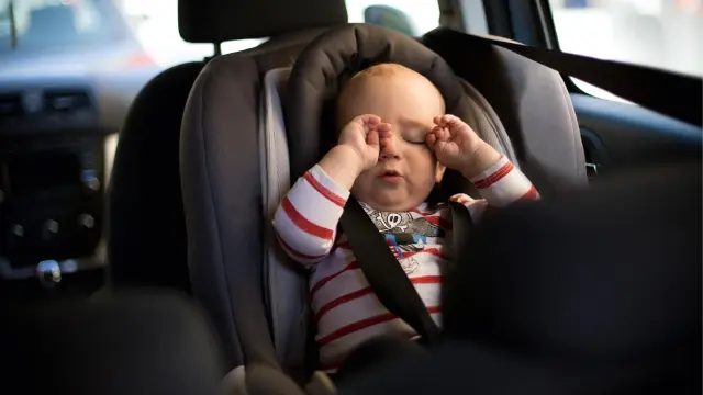 Ein Baby reibt sich die Augen, während es im Kindersitz eines Fahrzeuges sitzt. 