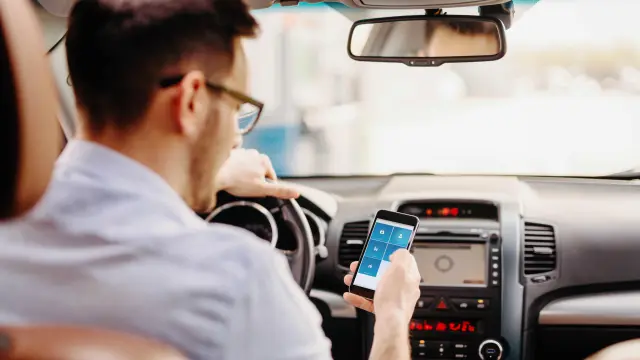 Un hombre se sienta en el asiento del conductor de su vehículo y mira su teléfono celular. 