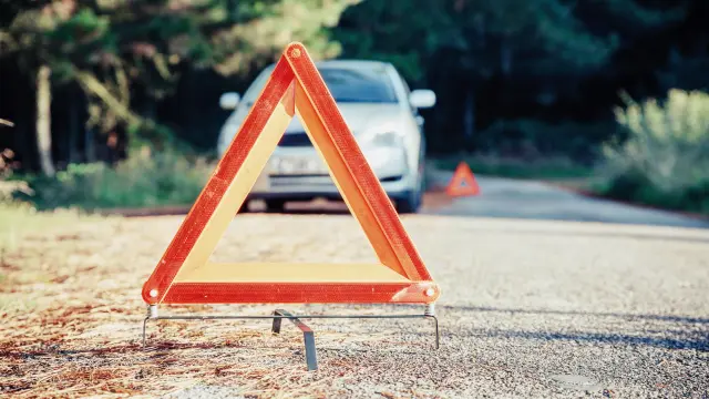 Ein Warndreieck steht auf einer Straße, im Hintergrund ist ein parkendes Fahrzeug zu sehen. 