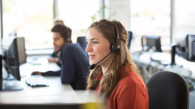 Una empleada del centro de servicios sonríe y mira la pantalla de su escritorio. Lleva una camiseta roja y utiliza unos auriculares para hacer llamadas telefónicas.