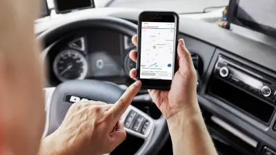 Ein Lkw-Fahrer sitzt im Führerhaus hinter dem Lenkrad und nutzt eine App auf seinem Smartphone