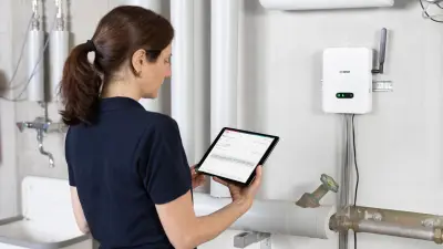 A technician stands in front of the heating system in a basement and looks at a tablet. The wall-mounted Anysight gateway can be seen in the background.