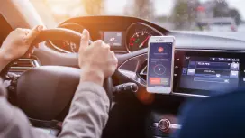 A person sits behind the steering wheel in a car with a smartphone visible on the center console.