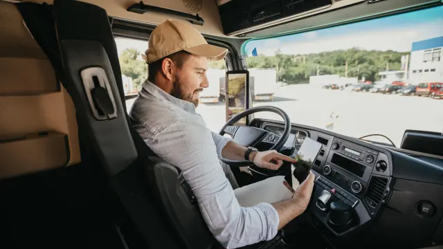 A truck driver sits in the cab of his truck and looks at a tablet.