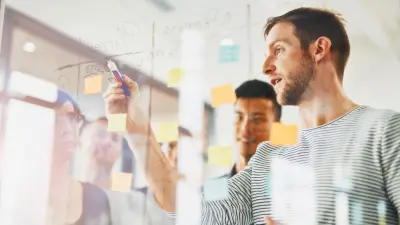 Un grupo de personas se encuentra frente a un metaplan transparente; una de ellas escribe algo en Post-Its con un rotulador permanente. 