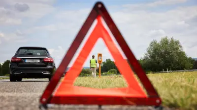 Blick durch ein Warndreieck auf eine Person, die gerade an einer Notrufsäule steht. Nebenan steht ein schwarzes Fahrzeug. 