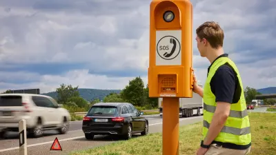 Eine Person steht vor einer Notrufsäule am Rande der Autobahn, er trägt eine gelbe Warnweste. Im Hintergrund ist ein schwarzer Fahrzeug auf dem Seitenstreifen zu sehen, dahinter steht ein Warndreieck. 