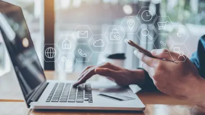A man sits in front of his laptop. In his left hand he is holding his smartphone, with his right hand he is handling his laptop. 