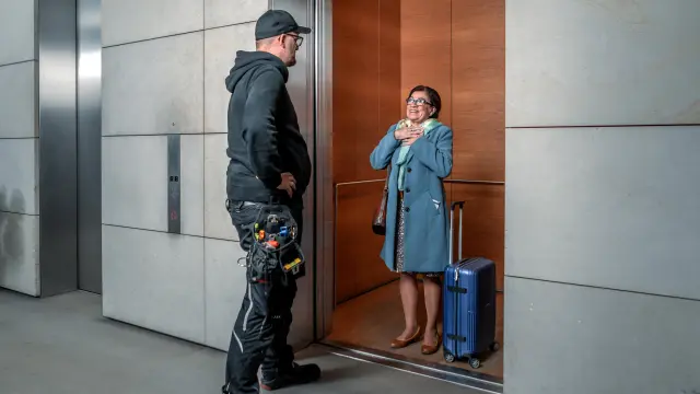 A relieved elderly lady looks out of the now open elevator door at the service technician.