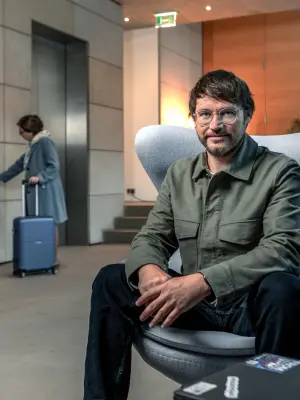 Sebastián Brix está sentado en un sillón y mira a la cámara; al fondo se ve a una señora delante de un ascensor.