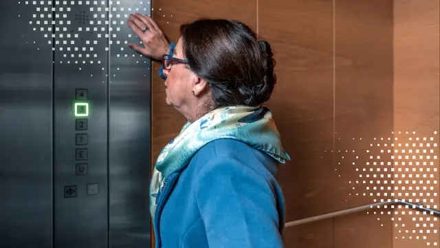 Eine ältere Dame steht in einem Aufzug und schaut auf das Display mit dem Notrufknopf. 