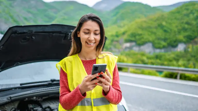 A woman is looking at her smartphone. She is wearing a yellow safety vest and is standing in front of her vehicle with the hood open.