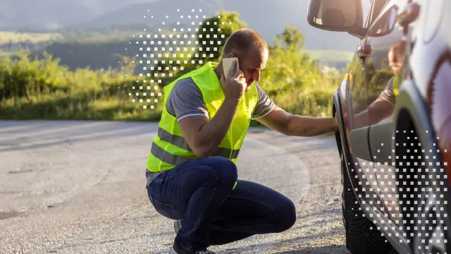 Ein Mann trägt eine gelbe Warnweste, er bückt sich neben seinem Wagen und schaut auf den Reifen seines Fahrzeugs. Er hat ein Smartphone am rechten Ohr und scheint zu telefonieren. 