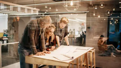 Three people are standing at a table, looking at a plan and making notes. The table is inside a glass room within an open space office with other people in the background.
