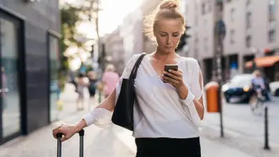 Eine blonde Frau schaut auf ihr Smartphone, welches sich in ihrer linken Hand befindet. Sie hat eine schwarze Tasche über der rechten Schulter und hält sich am Griff ihres Koffers fest. 