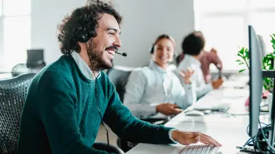 Ein Mitarbeiter im Service Center sitzt lächelnd vor seinem Bildschirm, er hat einen Schnurbart und trägt einen grünen Strickpullover. Im Hintergrund sind weitere Mitarbeitende zu sehen. 