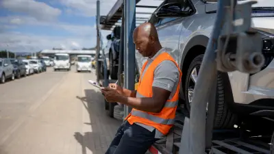 The driver of a car transporter is leaning against his truck trailer and writing something down on a clipboard. He is wearing an orange safety vest; other vehicles can be seen in the background.