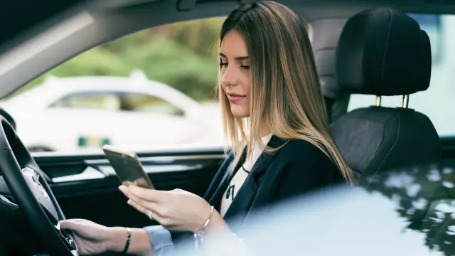 Eine Frau mit glatten langen Haaren sitzt in ihrem Fahrzeug und schau auf ihr Handy. 