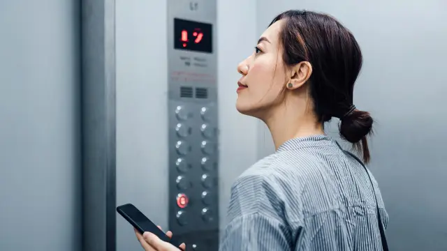 A lady is standing in an elevator, holding her smartphone.