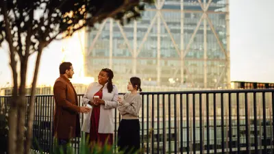 Drei Personen stehen miteinander auf einer Terrasse und unterhalten sich angeregt und lächelnd. Im Hintergrund ist ein hohes Gebäude in einer Stadt zu sehen. 