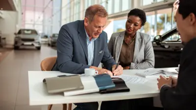 A woman and a man are sitting in front of a desk in a car dealership; the man is signing various agreement documents. 