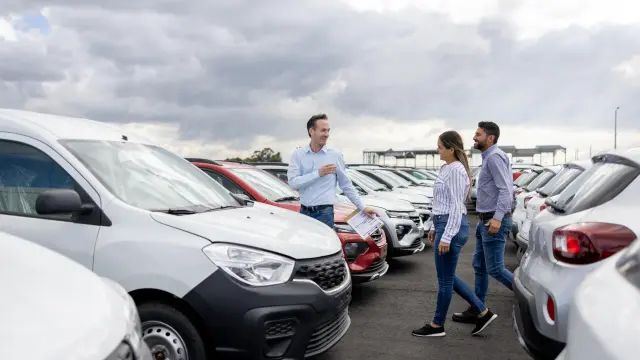 Two men and a woman are standing in a parking lot with many new cars.