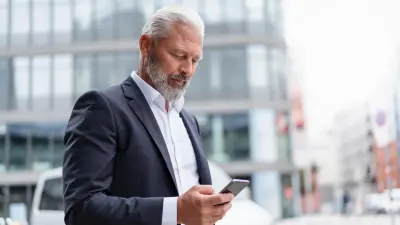 Un hombre de pelo canoso y con traje se para en la calle frente a un edificio y mira su smartphone.