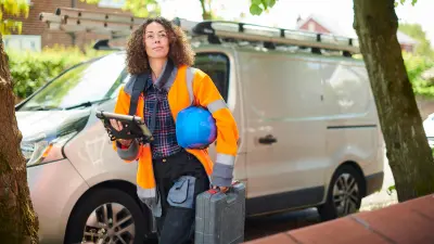 Una mujer de negocios se aleja de su vehículo con una tablet en la mano y llevando un casco azul bajo el brazo.
