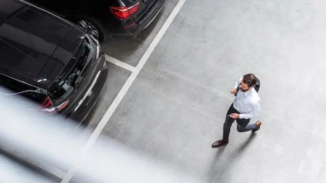 Mirando desde arriba se puede ver a un hombre corriendo a través de un garaje, hay algunos vehículos en negro. 