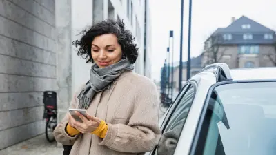 Una mujer con un abrigo de color beige está frente a su coche mirando el smartphone