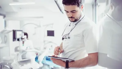 A dentist stands in a treatment room and looks at his tablet. Dentist chairs can be seen in the background.