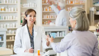 A pharmacist in a white coat stands behind the counter and accepts the credit card of her elderly customer. There are medicines between the two of them, and another pharmacist can be seen in the background.